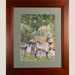 A framed watercolor painting of a garden scene with rocks, green foliage, and small orange and pink flowers, set in a mat and a warm brown wooden frame.