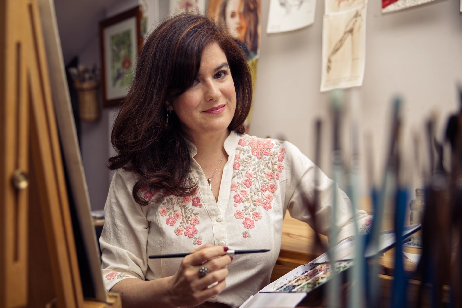 A woman with dark brown hair sits at an easel in an art studio, holding a paintbrush. She is wearing a cream-colored blouse with pink floral embroidery on the chest.